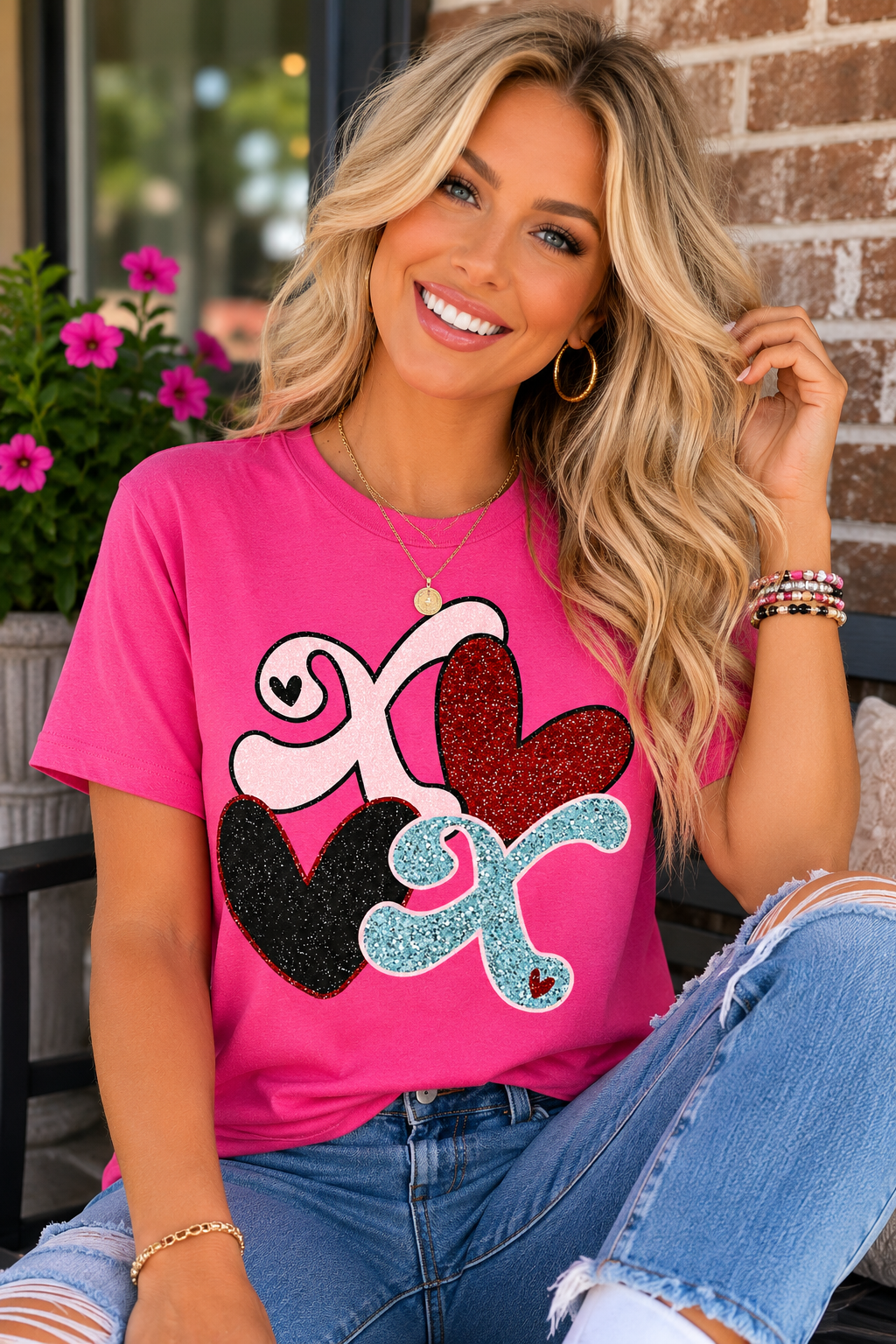 Woman wearing a bright pink graphic T-shirt featuring glitter-style XO letters and colorful hearts while smiling outdoors.
