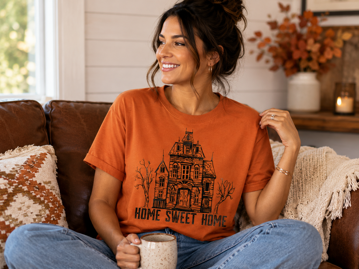 Woman wearing a burnt orange graphic T-shirt featuring a vintage haunted house and the words "Home Sweet Home" while relaxing indoors with a coffee mug.