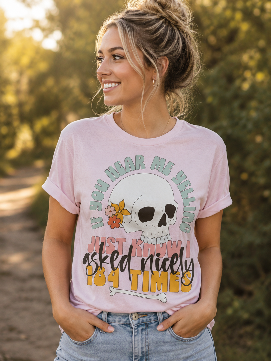 Woman wearing a light pink graphic T-shirt featuring a floral skull and the funny phrase "If You Hear Me Yelling I Already Asked Nicely 184 Times" while standing on a tree-lined path during golden hour.