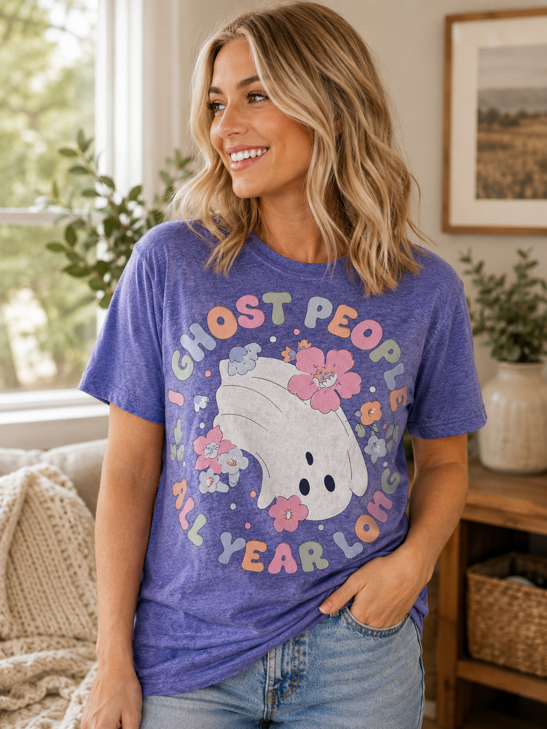 Woman wearing a purple graphic T-shirt featuring a cute floral ghost with the phrase "Ghost People All Year Long" while smiling indoors in a cozy home setting.