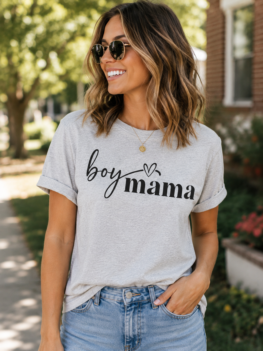 Smiling woman wearing a light heather gray "Boy Mama" graphic T-shirt with black script lettering while walking outdoors in sunglasses and blue jeans.