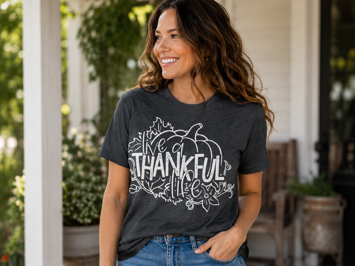 Woman wearing a heather charcoal gray T-shirt featuring a white pumpkin graphic with the phrase "Live a Thankful Life" surrounded by autumn leaves and floral accents while smiling on a front porch.