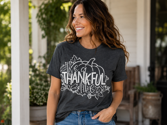 Woman wearing a heather charcoal gray T-shirt featuring a white pumpkin graphic with the phrase "Live a Thankful Life" surrounded by autumn leaves and floral accents while smiling on a front porch.