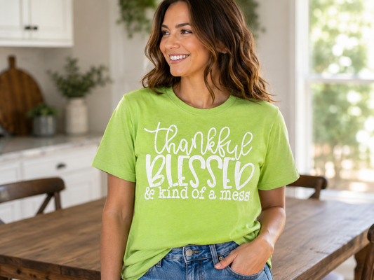 Woman wearing a bright lime green T-shirt featuring the phrase "Thankful, Blessed & Kind of a Mess" in white lettering while smiling in a bright kitchen.
