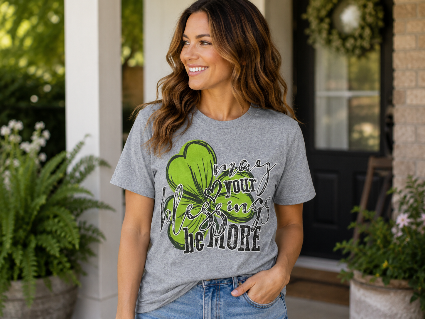 Woman wearing a heather gray T-shirt featuring a green shamrock graphic with the phrase "May Your Blessings Be More" while smiling on a front porch.