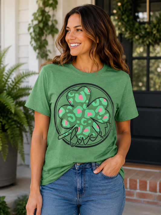 Woman wearing a heather green T-shirt featuring a green and pink leopard print shamrock with the word "Lucky" in black script while smiling on a front porch.