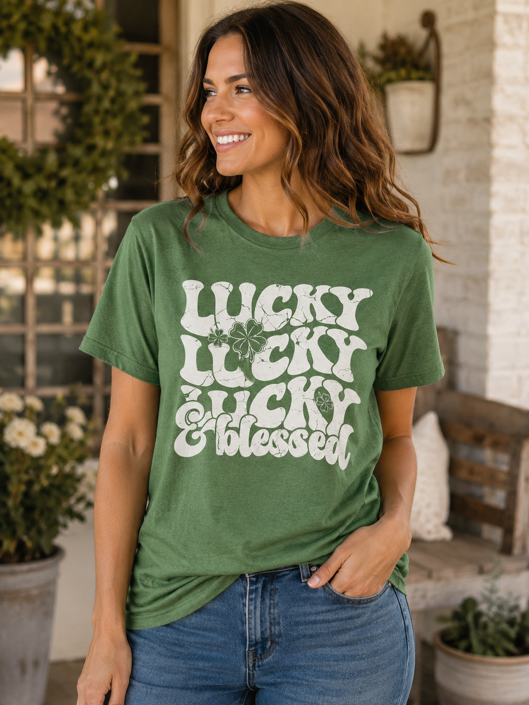 Woman wearing a heather green graphic T-shirt featuring the words "Lucky Lucky Lucky & Blessed" with shamrock accents while smiling on a front porch.