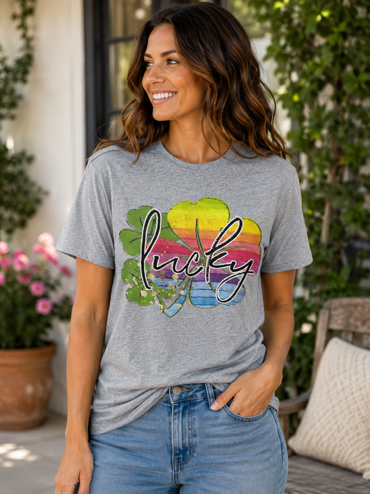Woman wearing a heather gray graphic T-shirt featuring a colorful rainbow shamrock with the word "Lucky" while smiling outdoors on a front porch.