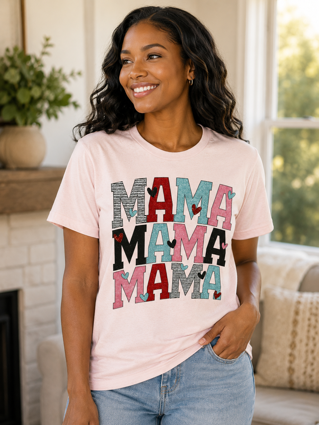 Woman wearing a light pink graphic T-shirt featuring a colorful stacked "Mama Mama Mama" design with heart accents while smiling indoors.