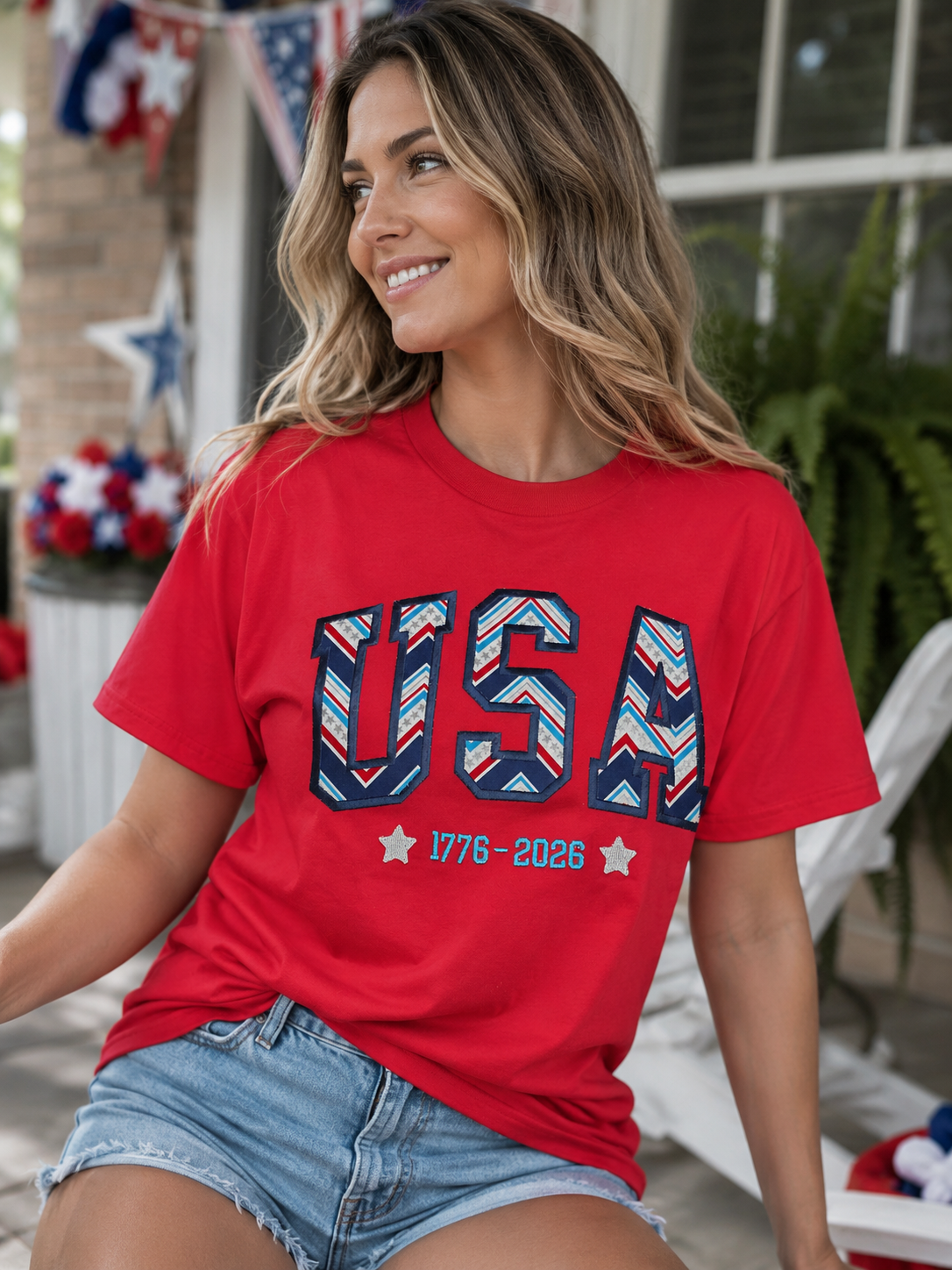 Woman wearing a red USA 1776–2026 patriotic graphic T-shirt featuring red, white, and blue varsity lettering while sitting outdoors with Fourth of July decorations in the background.