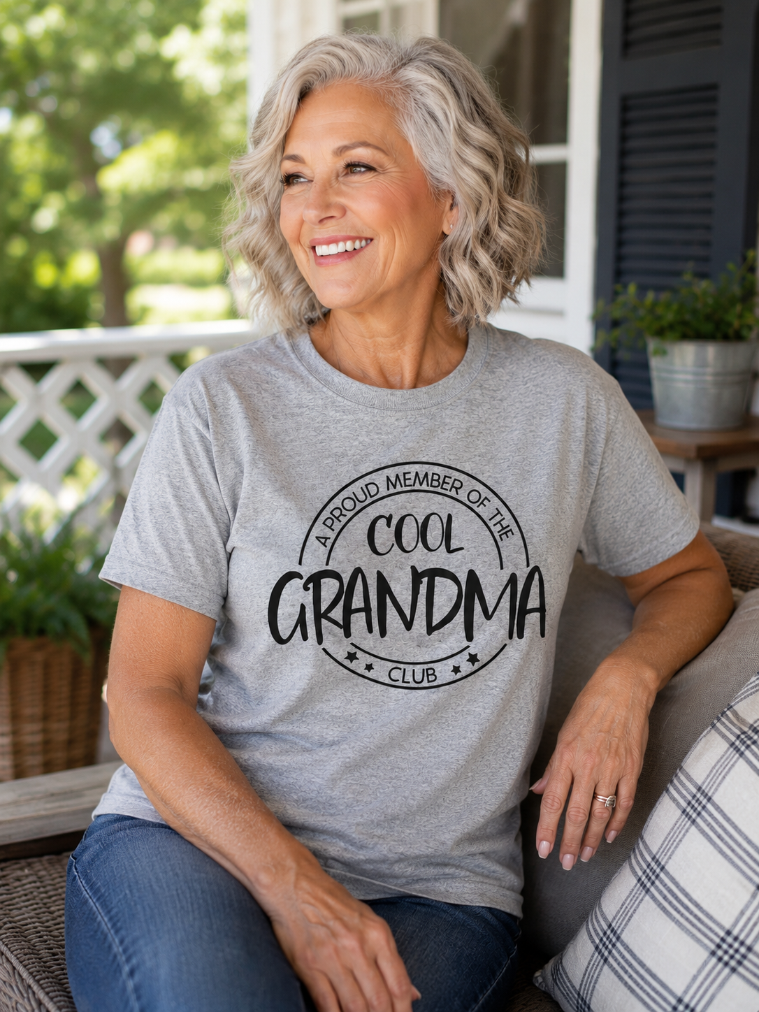 Smiling woman wearing a heather gray T-shirt featuring the words "A Proud Member of the Cool Grandma Club" while relaxing on a front porch.