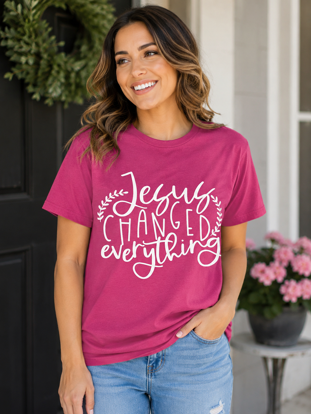Woman wearing a berry pink T-shirt featuring a white "Jesus Changed Everything" Christian graphic while standing on a front porch with flowers in the background.
