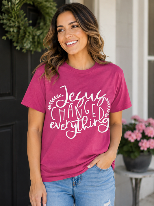 Woman wearing a berry pink T-shirt featuring a white "Jesus Changed Everything" Christian graphic while standing on a front porch with flowers in the background.