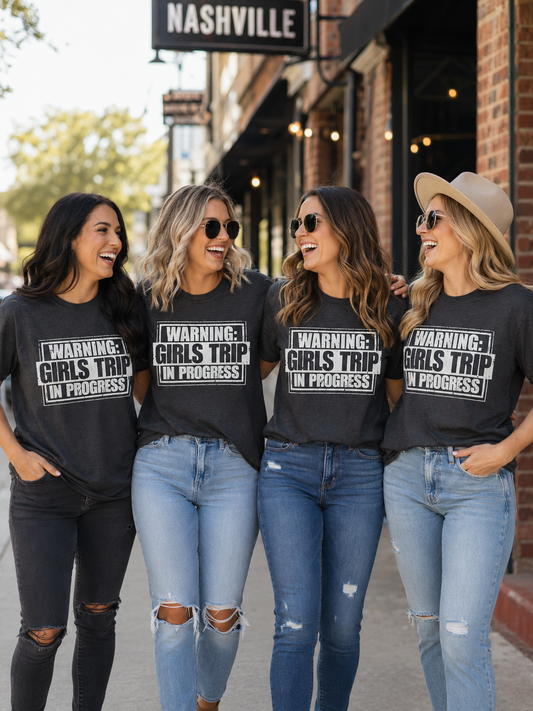 Four women wearing matching charcoal gray graphic T-shirts that read "Warning: Girls Trip in Progress" while walking together and laughing on a downtown street in Nashville.