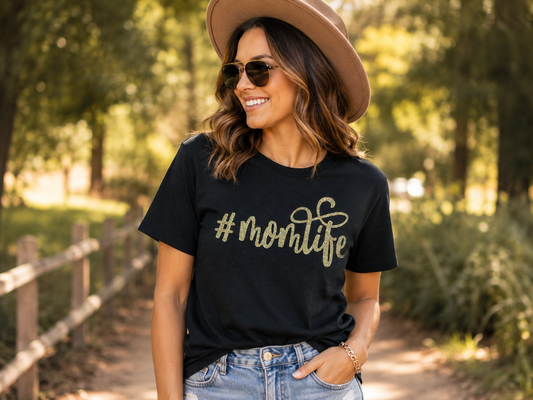 Woman wearing a black T-shirt with a gold "#MomLife" script graphic while standing on a tree-lined path wearing a tan hat and sunglasses.