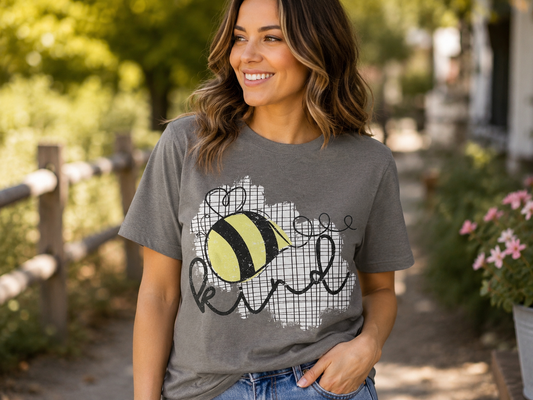 Woman wearing a heather charcoal gray T-shirt featuring a yellow and black honey bee graphic with the word "Kind" while smiling outdoors on a sunny path.